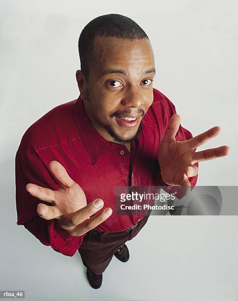 handsome young african american adult male with facial hair wearing a red shirt stands looking up at the camera as he gestures with his hands and gives a quizzical facial expression - african male red shirt stock-fotos und bilder