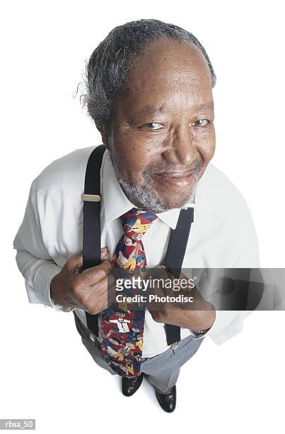 attractive old african american adult male with facial hair and balding gray hair wearing a white dress shirt and a humorous tie and suspenders stands looking up at the camera with his hands on his chest holding his suspenders while smiling - white shirt ストックフォトと画像