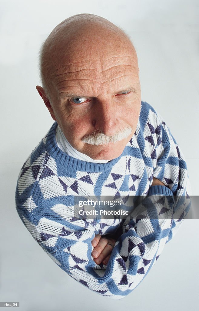 Handsome blue eyed old bald caucasian adult male with a moustache wearing a blue and white patterned sweater stands with arms folded looking up at the camera with one eye staring and an accusative expression on his face