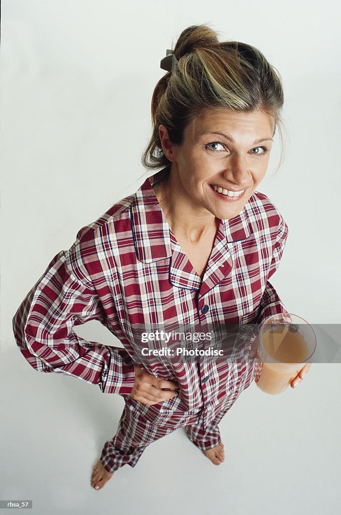 Pretty young adult blonde caucasian female with her hair up is dressed in checkered red pajamasand is barefoot as she stands holding a glass of orange juice and smiles up at the camera humorously