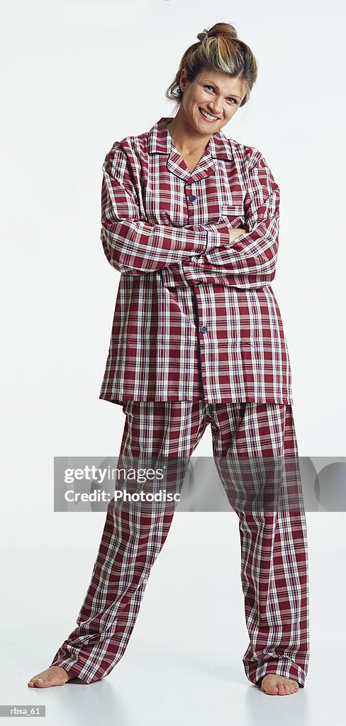 Pretty young caucasian adult blonde female barefoot with hair up wearing red plaid pajamas stands with arms folded looking at the camera with a humorous smile
