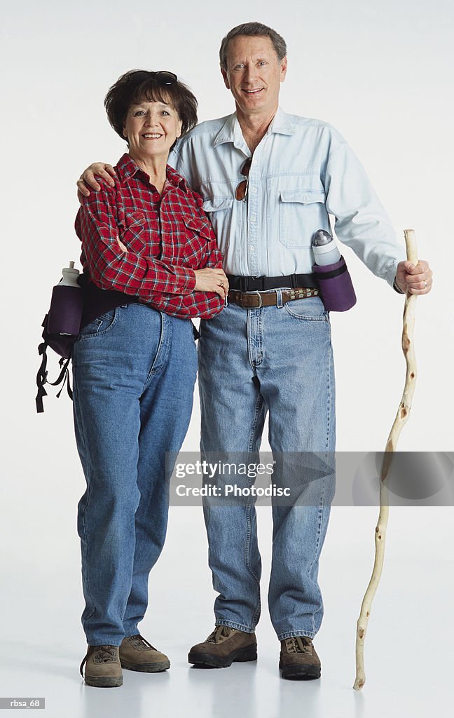 Middle aged adult caucasian female wearing jeans and a red flannel shirt with a water bottle standing with middle aged adult caucasian male wearing jeans and a blue shirt and water bottle holding hiking stick and looking at the camera smiling as a co
