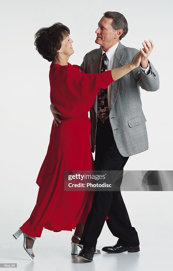 Middle aged adult caucasian brunette female wearing a long red dress and middle aged adult caucasian gray haired male wearing a suit and tie holding each other in a dance pose as they look into each others eyes smiling