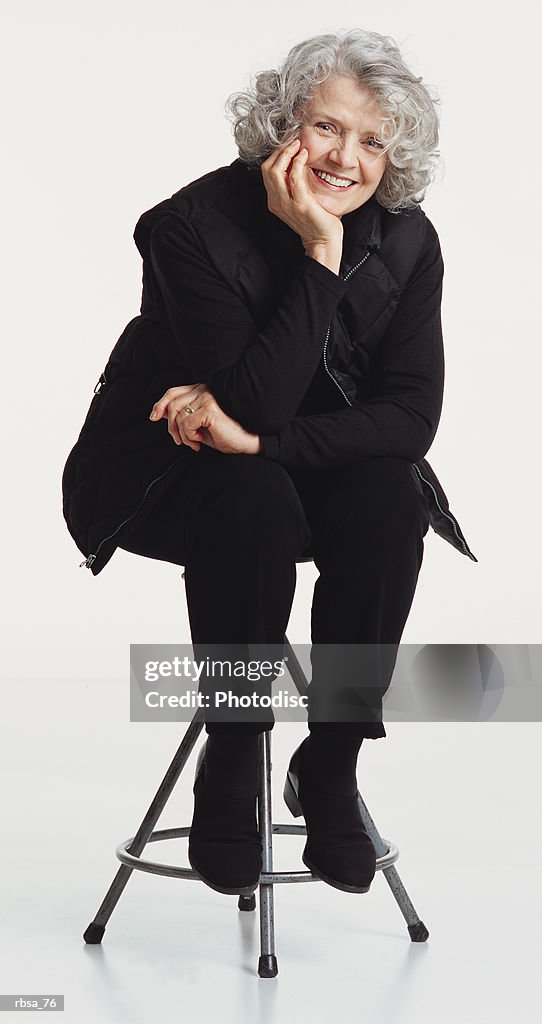 Pretty old caucasian adult female with curly gray hair wearing a dark vest and dark pants sitting on a stool as she leans forward resting her chin in her hand and smiles at the camera