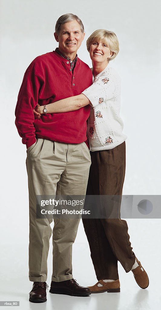 Old caucasian adult male with gray hair wearing a red sweater and tan slacks stands while old caucasian adult female with blonde hair wearing a white sweater and brown slacks stands alongside him with her arms around his waist as the couple smiles at
