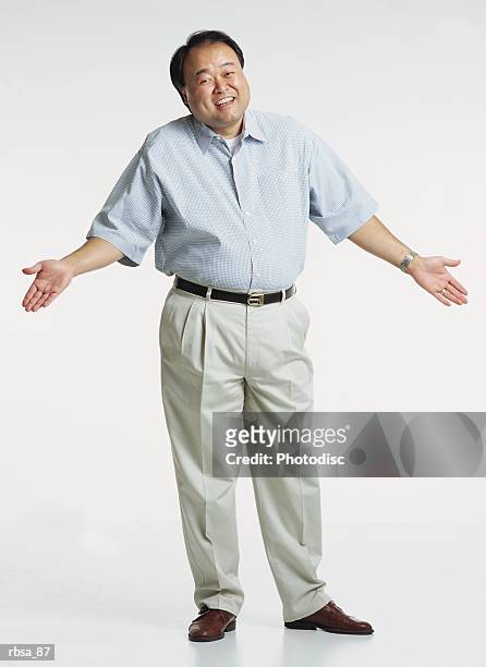 handsome middle aged asian adult male wearing a light blue short sleeved shirt and cream colored slacks stands with shrugged shoulders and hands in the air as he looks at the camera with a anxious smile - smile stockfoto's en -beelden