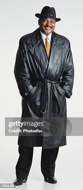 handsome middle aged adult african american male with a moustache and hat wears a dark leather trench coat and white shirt and tie while standing and looking at the camera smiling approvingly - gabardina larga fotografías e imágenes de stock