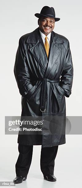 handsome middle aged adult african american male with a moustache and hat wears a dark leather trench coat and white shirt and tie while standing and looking at the camera smiling approvingly - trench coat stock pictures, royalty-free photos & images