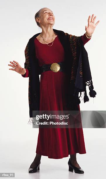 old stylish caucasian gray haired adult female wearing a red dress with a large belt buckle and a colorful shawl  stands posing with her head tilted back and hands raised slightly in a gesture of drama as she her body faces the camera and her gaze is - belt stockfoto's en -beelden