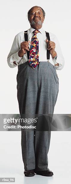 old african american adult male with gray hair and facial hair wearing  a white shirt and bright tie with gray slacks and suspenders stands alertly while holding his suspenders away from his chest and looking at the camera with a humorous smile and t - smile stockfoto's en -beelden
