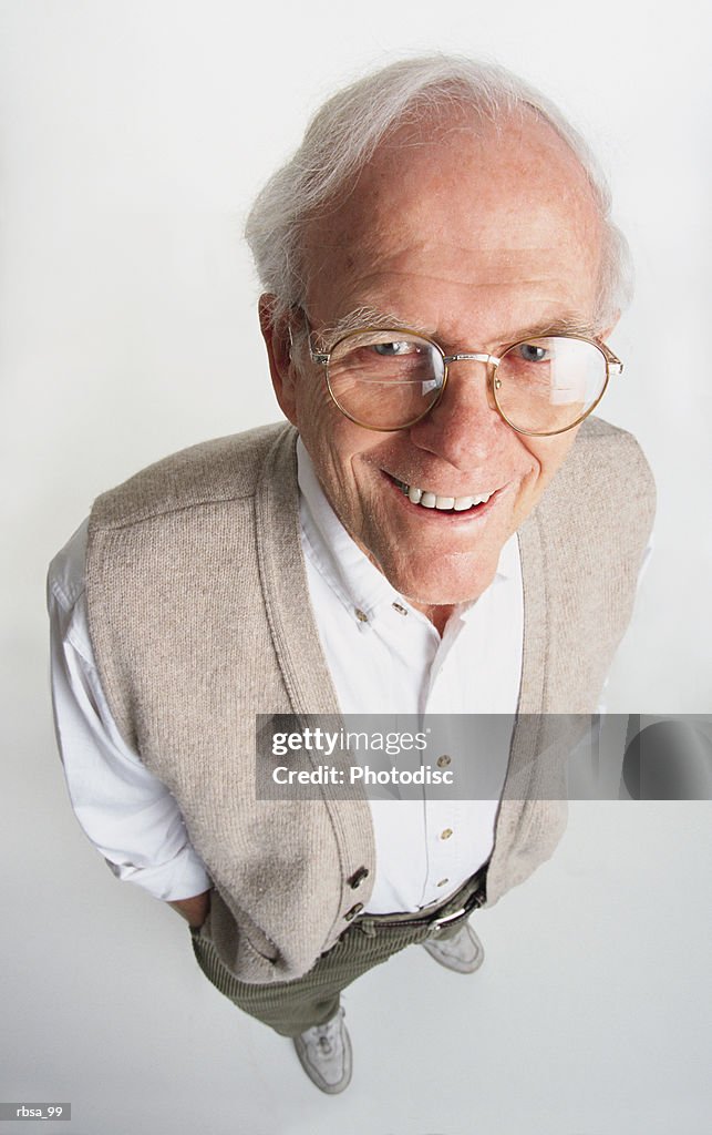 Attractive old adult male caucasian with white hair and glasses wearing a tan vest and white shirt stands while looking up at the camera with a kind smile