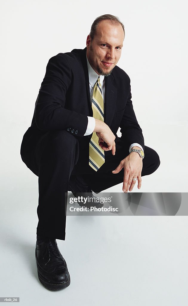 A handsome young caucasian male in a dark business suit is crouched down on his back leg looking into the camera