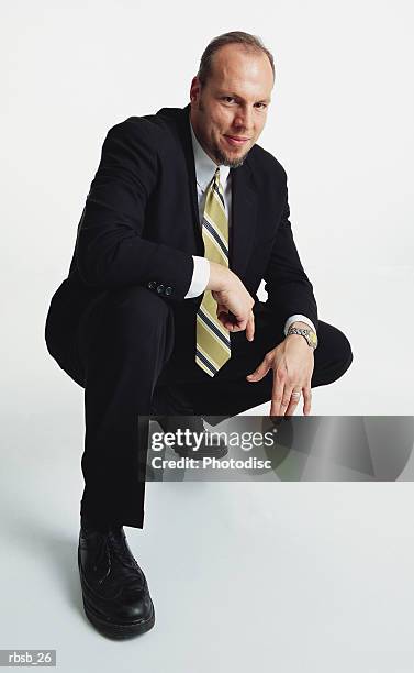 a handsome young caucasian male in a dark business suit is crouched down on his back leg looking into the camera - male portrait suit and tie stock-fotos und bilder