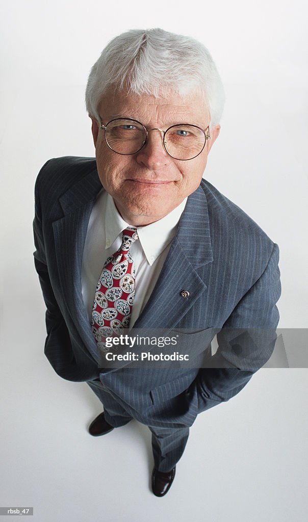 A handsome middle aged caucasian businessman with glasses and white hair is dressed in a grey suit with his hand in his pockets and looking up at the camera