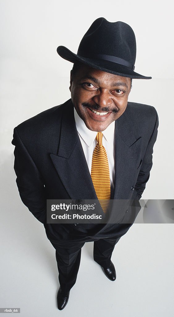 A handsome middle aged african american man with black hat and dark business suit looking up into the camera with his hands in his pockets