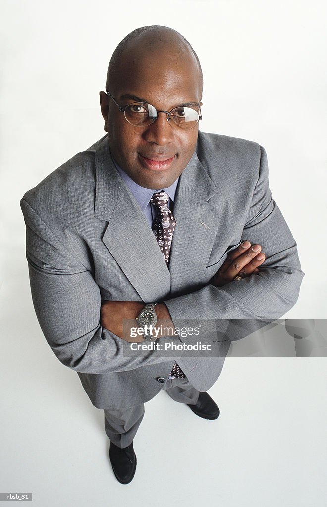 A handsome middle aged bald african american man in a light grey business suit stands looking up at the camera with his arms crossed
