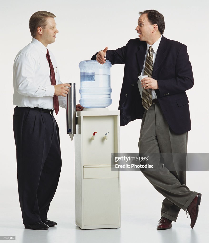 Two young caucasian businessmen are standing around a watercooler conversing with eachother