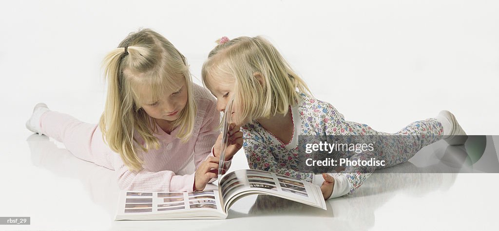 Two blonde sisters in thermals/pajamas read a book together while they lay on their stomachs talking