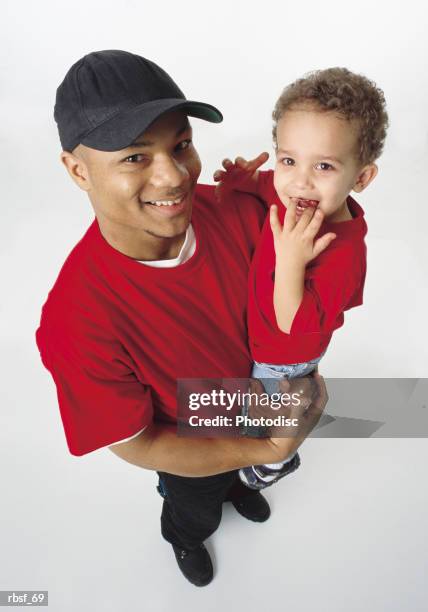 african american single father holds young son and smiles wearing baseball cap and red shirt - genderblend stock-fotos und bilder
