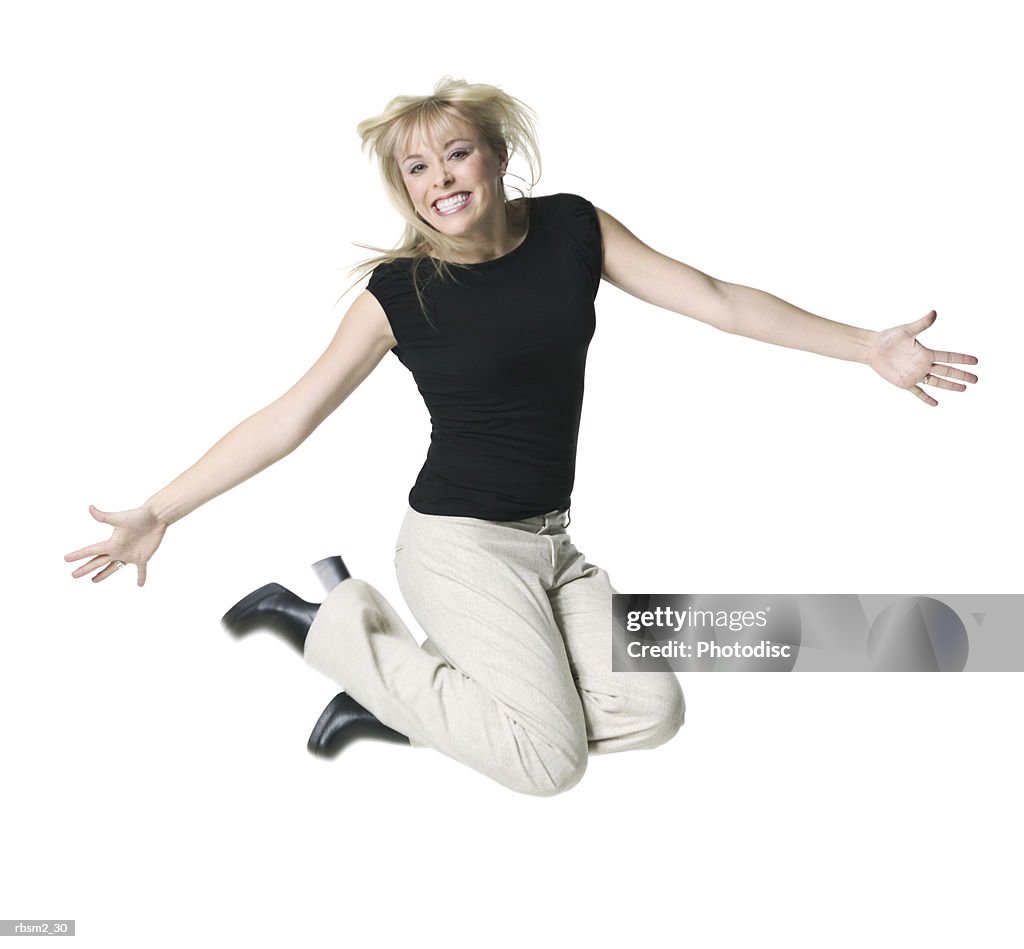 A young adult blonde female in tan pants and a black shirt jumps up and smiles