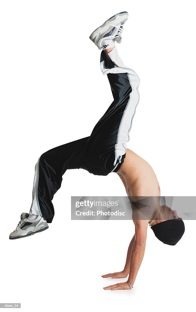 A young caucasian male breakdancer in black pants and no shirt and a knit cap is flipped upside down balancing on his hands with his legs in the air