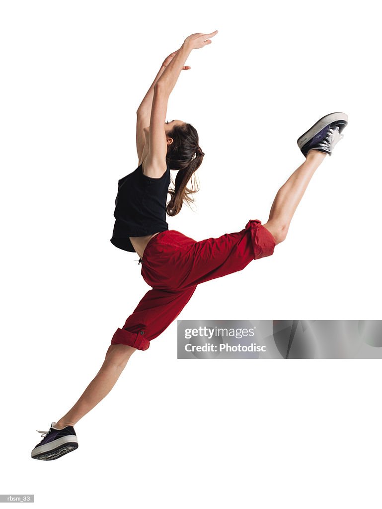 A teenage caucasian female dancer in red pants and a black tank top jumps forward raising her arms and throwing her leg back