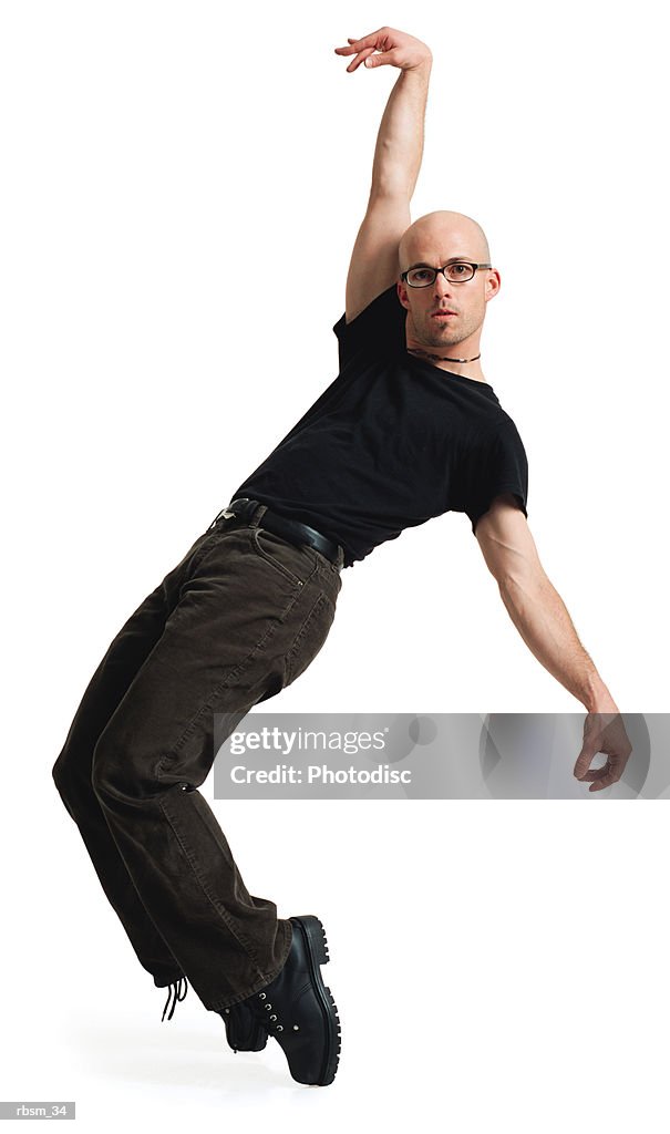 A young caucasian male modern dancer with a bald head in glasses black pants and t-shirt jumps up on heis toes arn raises one arm