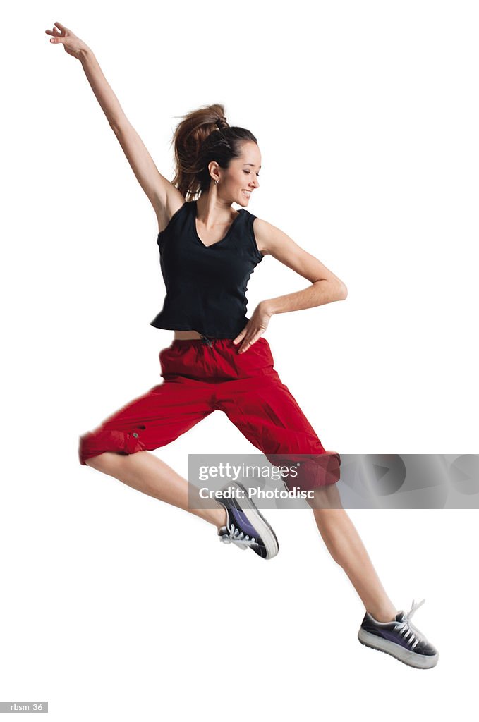 A teenage caucasian female dancer in red pants and a black tank top jumps up and to the side while raising one arm