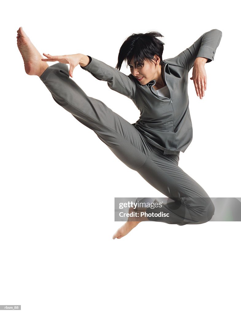 A young latin female dancer in a grey outfit jumps into the air and kicks in a karate type pose