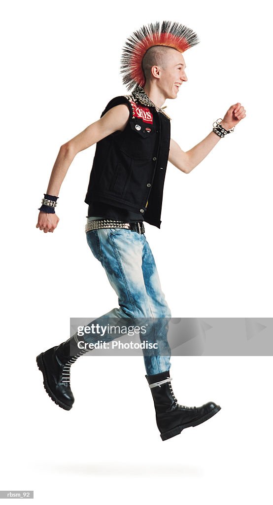 A young caucasian male punker with a colorful mohawk in jeans and black vest jumps up and smiles