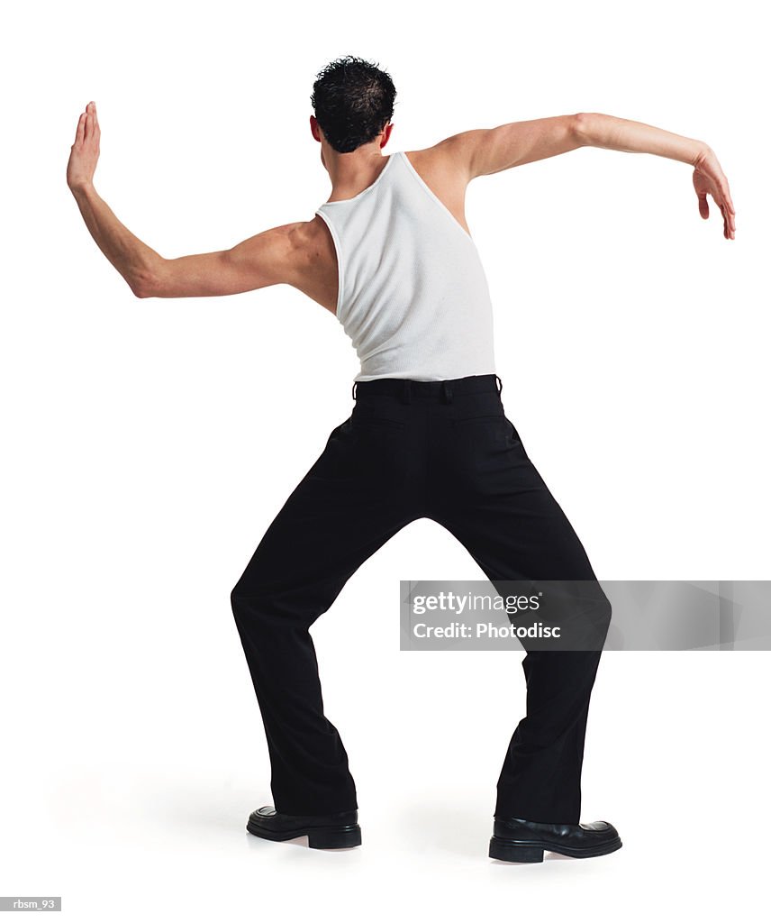A young caucasian male dancer in black pants and white tank top stretches out and moves his arms with his back to the camera