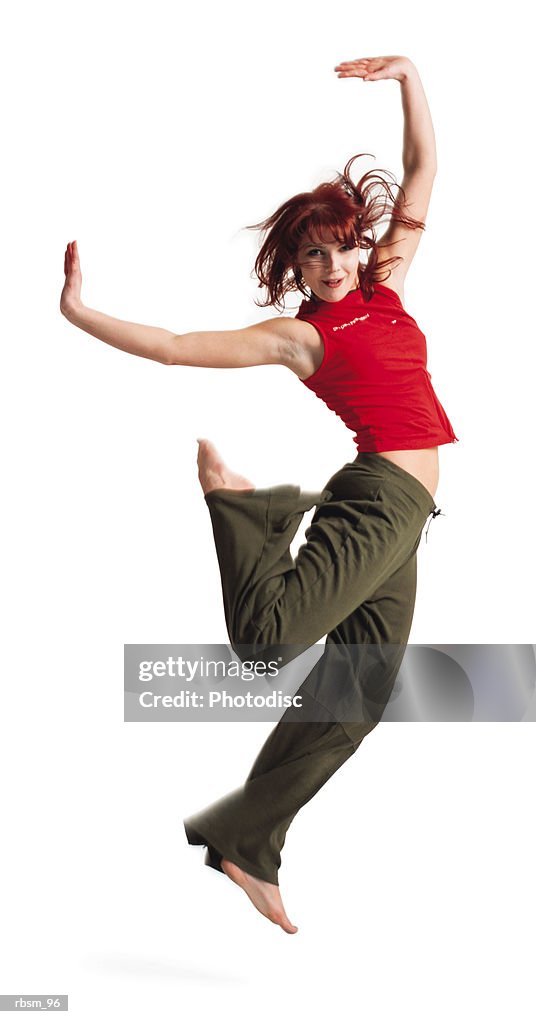 An attractive caucasian female in green pants and a red tank top hops up into the air as she throws her arms up