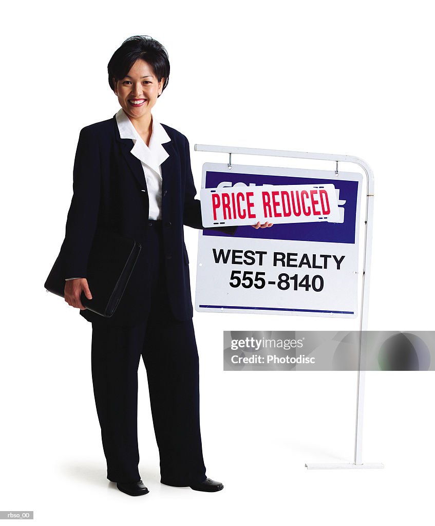 An asian female realator stands smiling by a for sale sign