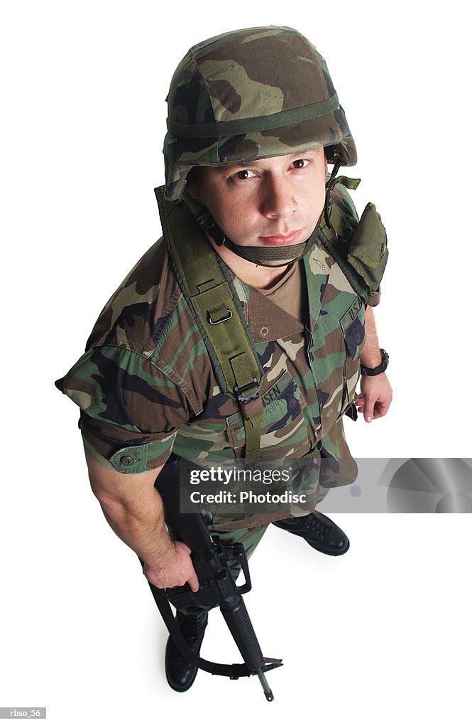 A caucasian male soldier in fatigues and holding a gun looks sternly up at the camera