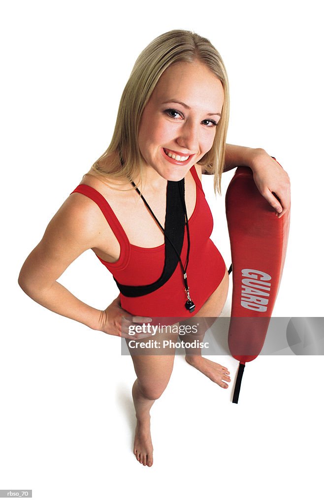 A blonde female lifeguard in a red swimsuit stands with her rescue tube as she smiles and looks up at the camera