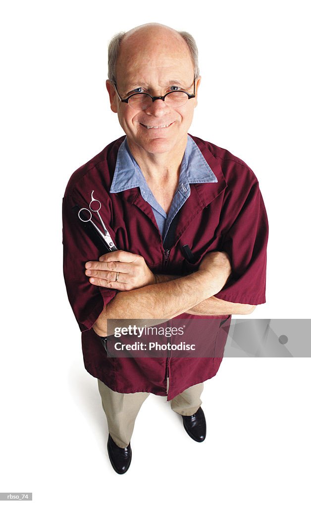 An elderly caucasian barber folds his arms and holds his scissors as he looks up at the camera