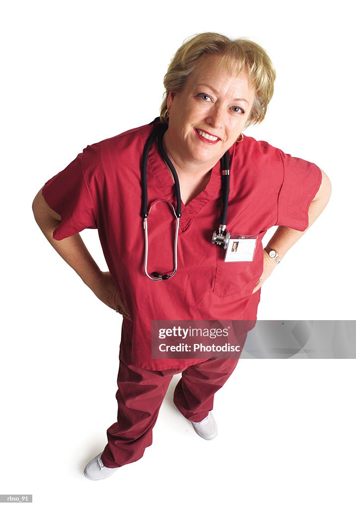 An older caucasian female nurse dressed in pink scubs places her hands on her hips and looks up at the camera