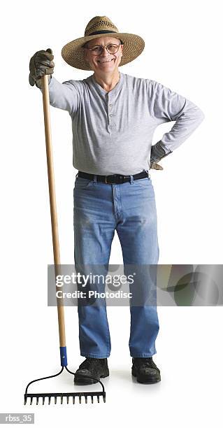 elderly man wearing a straw hat and holding a rake smiles at the camera. - rake stock-fotos und bilder