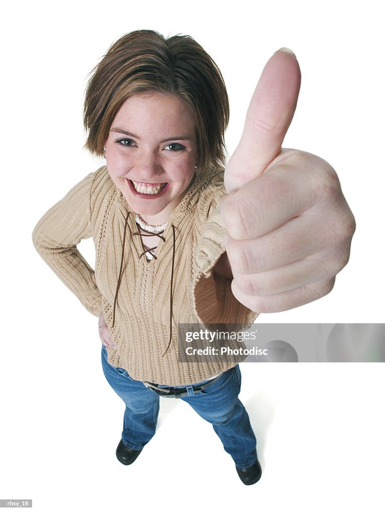 A caucasian teenage girl in jeans and a tan sweater gives the thumbs up sign up into the camera