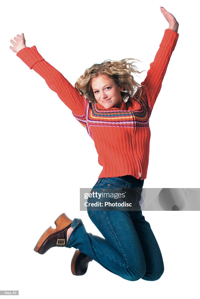 Caucasian blonde female teen in jeans and red sweater jumps up into the air and throws her arms up
