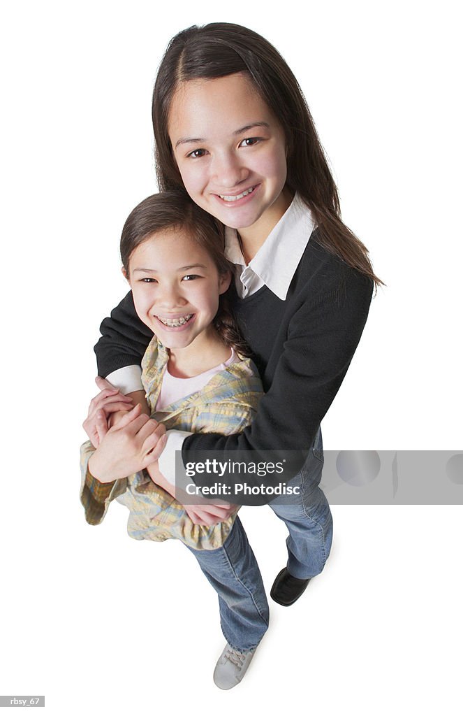 An asian teenage girl in jeans and a black sweater wraps her arms around her little sister and smiles up into the camera