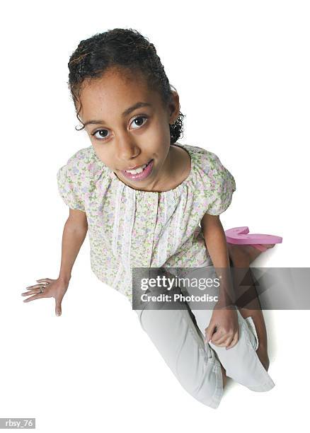 a young african american girl in tan pants and a green floral shirt sits and smiles up into the camera - tan tan stock pictures, royalty-free photos & images