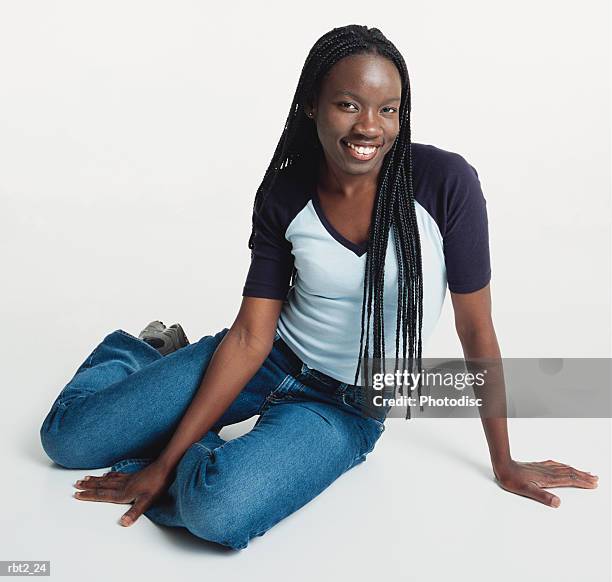 a beautiful young black woman with long cornrows sitting sideways on the floor wearing jeans and a teeshirt smiles at the camera - braided hairstyles for african american girls stock pictures, royalty-free photos & images