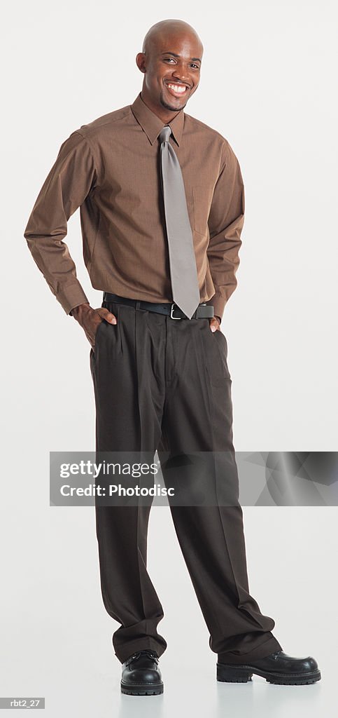 A handsome bald black young man with a goatee wearing slacks and a brown dress shirt and ties stands with hands in pockets and smiles at the camera