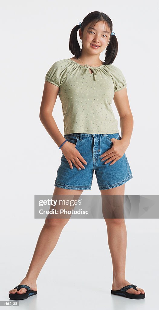 A cute asian girl in ponytails and blue shorts stands with her thumbs in her pockets and smiles at the camera