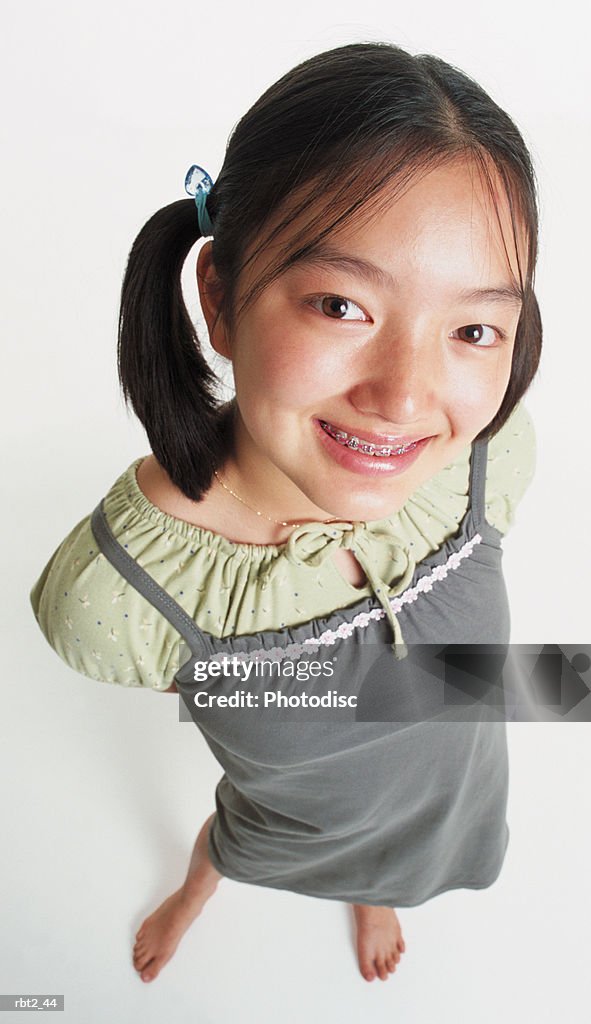 A cute asian girl in ponytails and braces wears a gray jumper and stands barefooted looking up into the camera