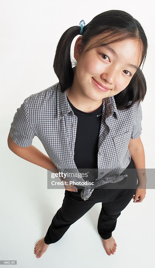 A cute young asian girl with ponytails wearing black stretch pants with a checkered blouse looks up at the camera smiling with her head cocked