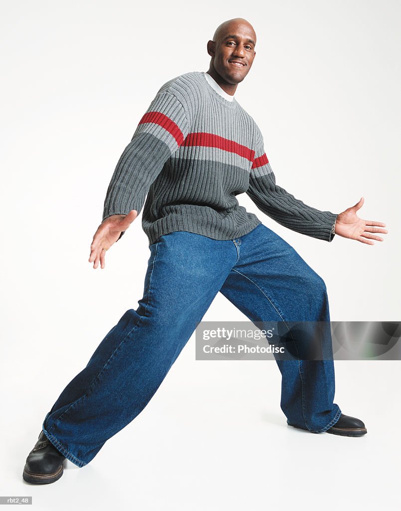 A bald young african american man in baggy jeans and gray sweater strikes an animated pose with arms and legs spread wide while looking toward the camera