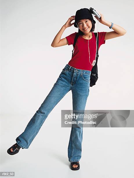 a pretty asian preteen girl wearing a red teeshirt and blue jeans has a black hat on her head and is listening to a walkman and wearing a backpack while standing on one foot - personal compact disc player 個照片及圖片檔