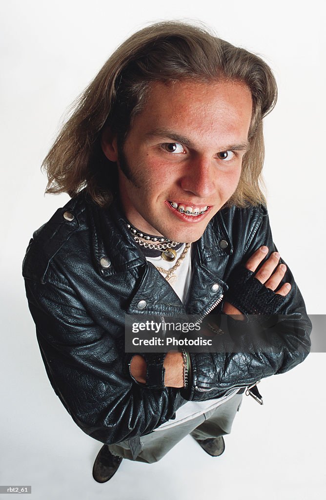 A teenage caucasian boy with long hair and braces is wearing choke chains and a leather jacket as he folds his arms and smiles up at the camera